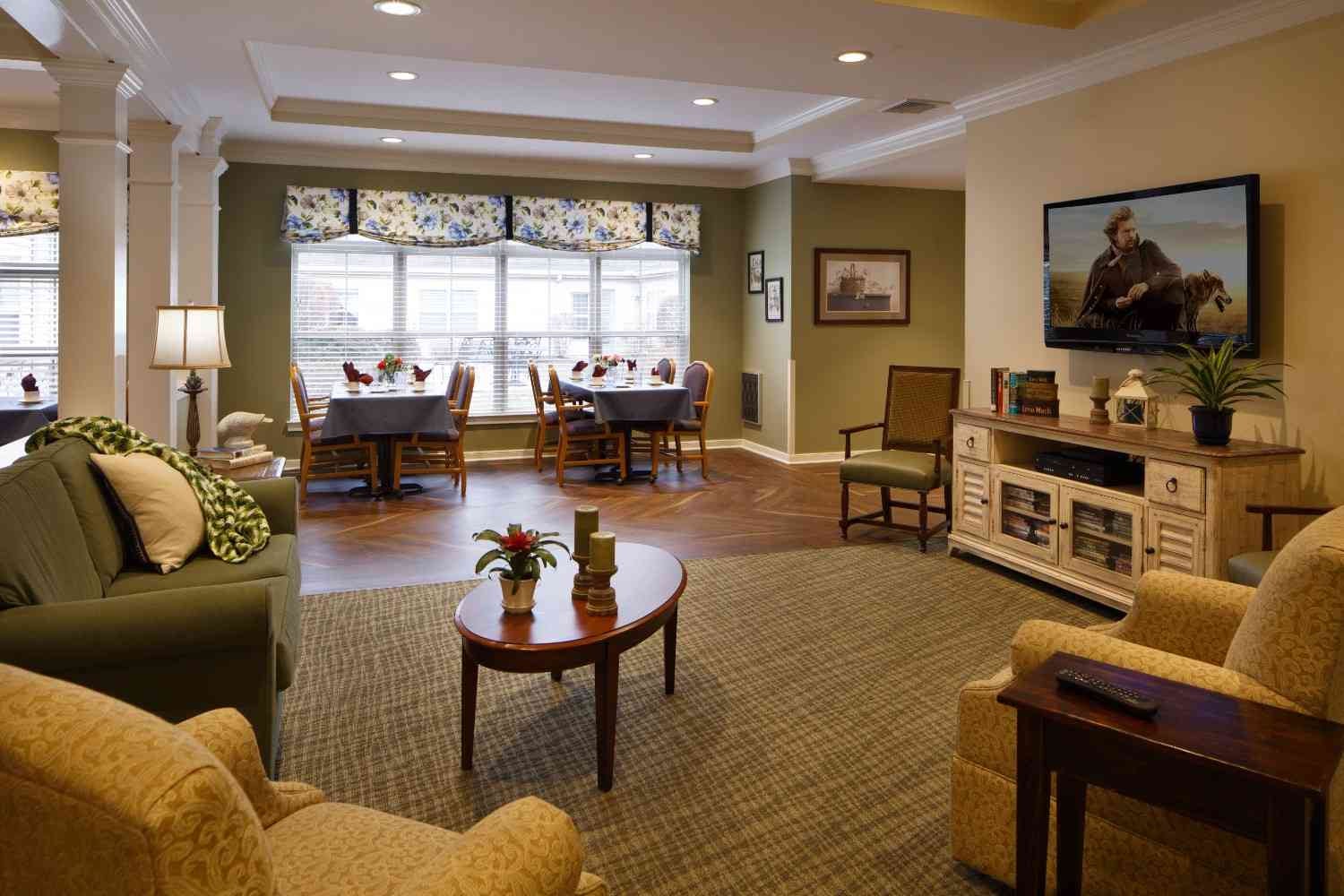 A brightly lit living room features a television, seating area, and dining table with floral accents.